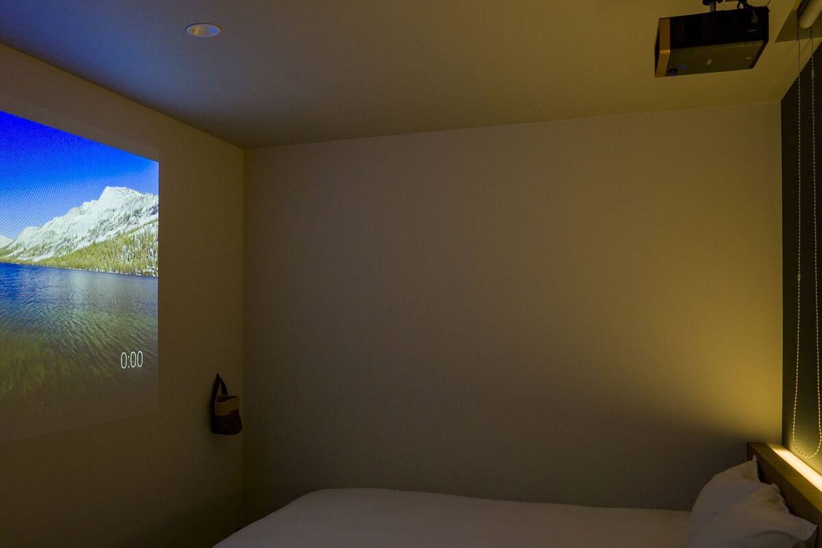 Ein Hotelzimmer mit einem Bett, einem an der Wand befestigten Projektor, der ein Landschaftsbild zeigt, und einem kleinen Nachttisch mit einer Lampe.