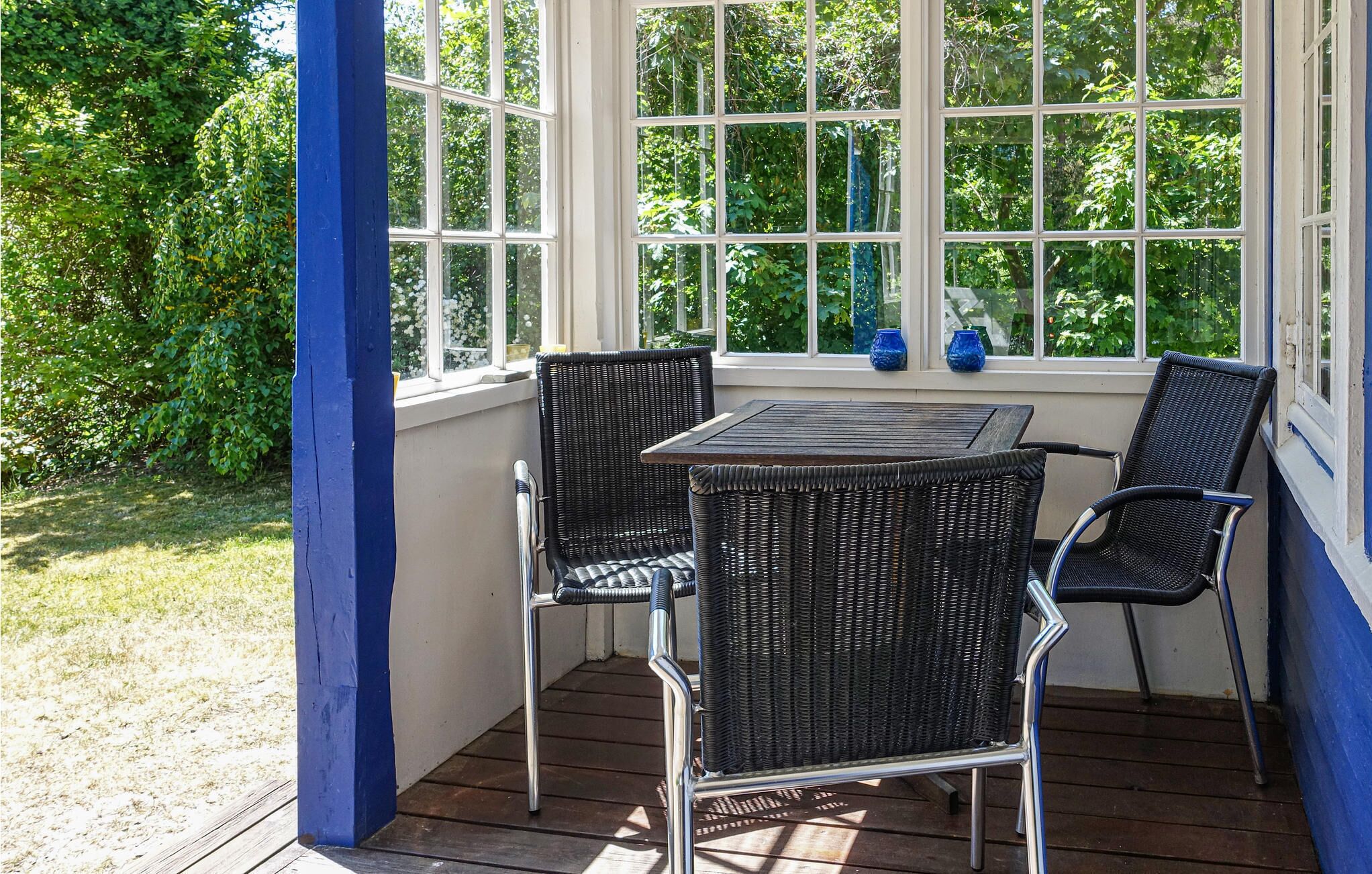 A covered patio with a wooden table and four wicker chairs, large windows, and a view of greenery.
