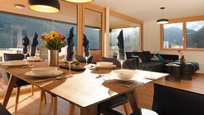 Un espace repas moderne avec une table en bois dressée pour quatre personnes, de grandes fenêtres offrant une vue sur les montagnes et un coin salon confortable avec un canapé et des tables d’appoint.