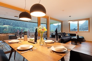 A modern dining room with a wooden table set for a meal, large windows offering a mountain view, and a cozy seating area with a sofa and side table.
