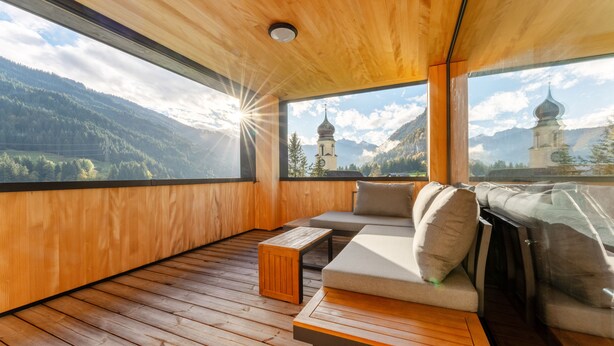 Une terrasse en bois avec un canapé et une petite table, offrant une vue sur un paysage montagneux avec une église.