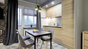 A modern kitchen with a dining area, featuring a table with chairs, a fruit bowl, and a window with curtains.