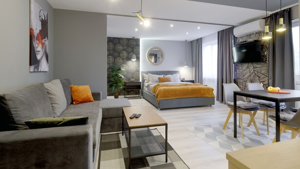 A modern hotel room with a grey sofa, a bed with a yellow blanket, a dining table with chairs, and a TV on the wall.
