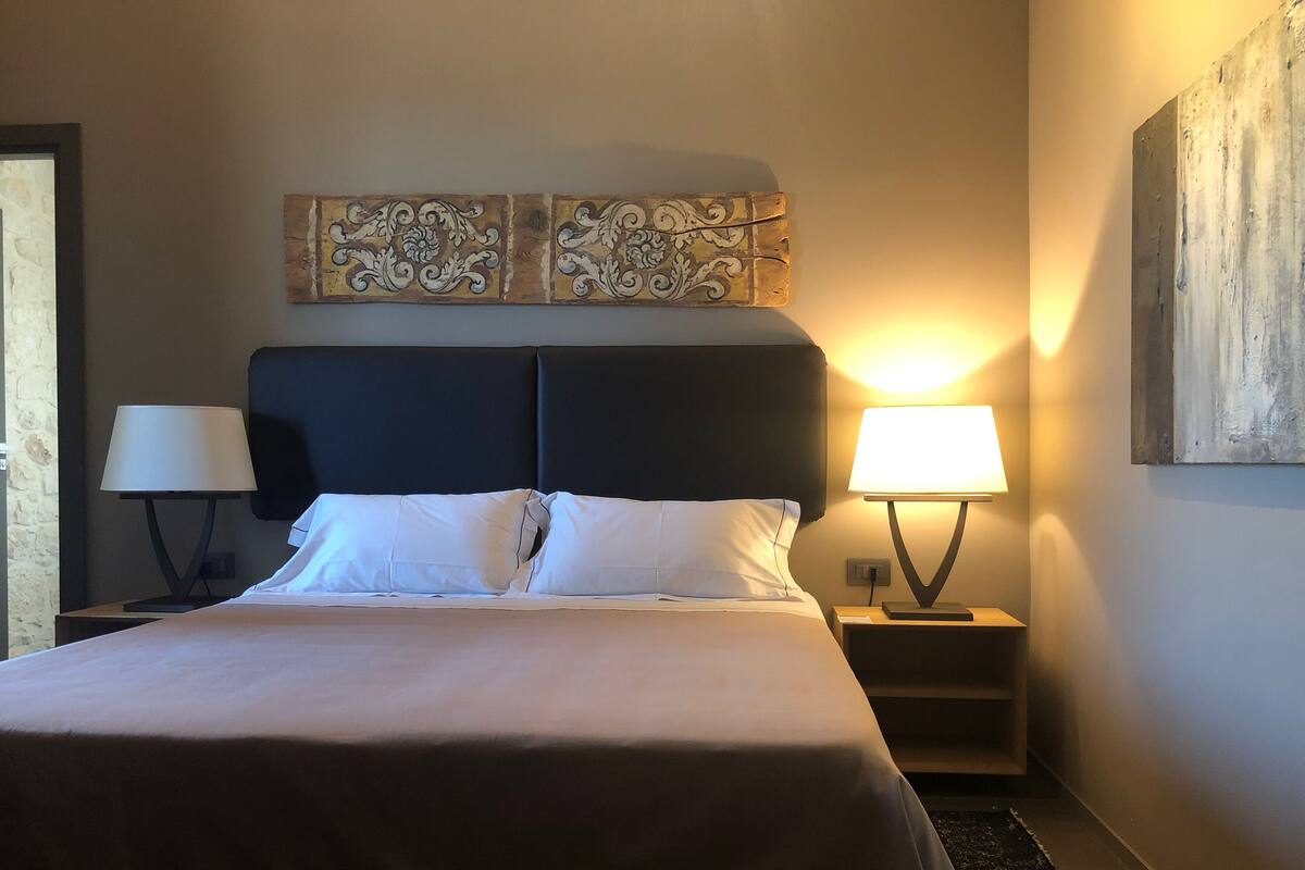 A neatly made bed with white linens, two bedside lamps, a decorative headboard, and a wall-mounted artwork.