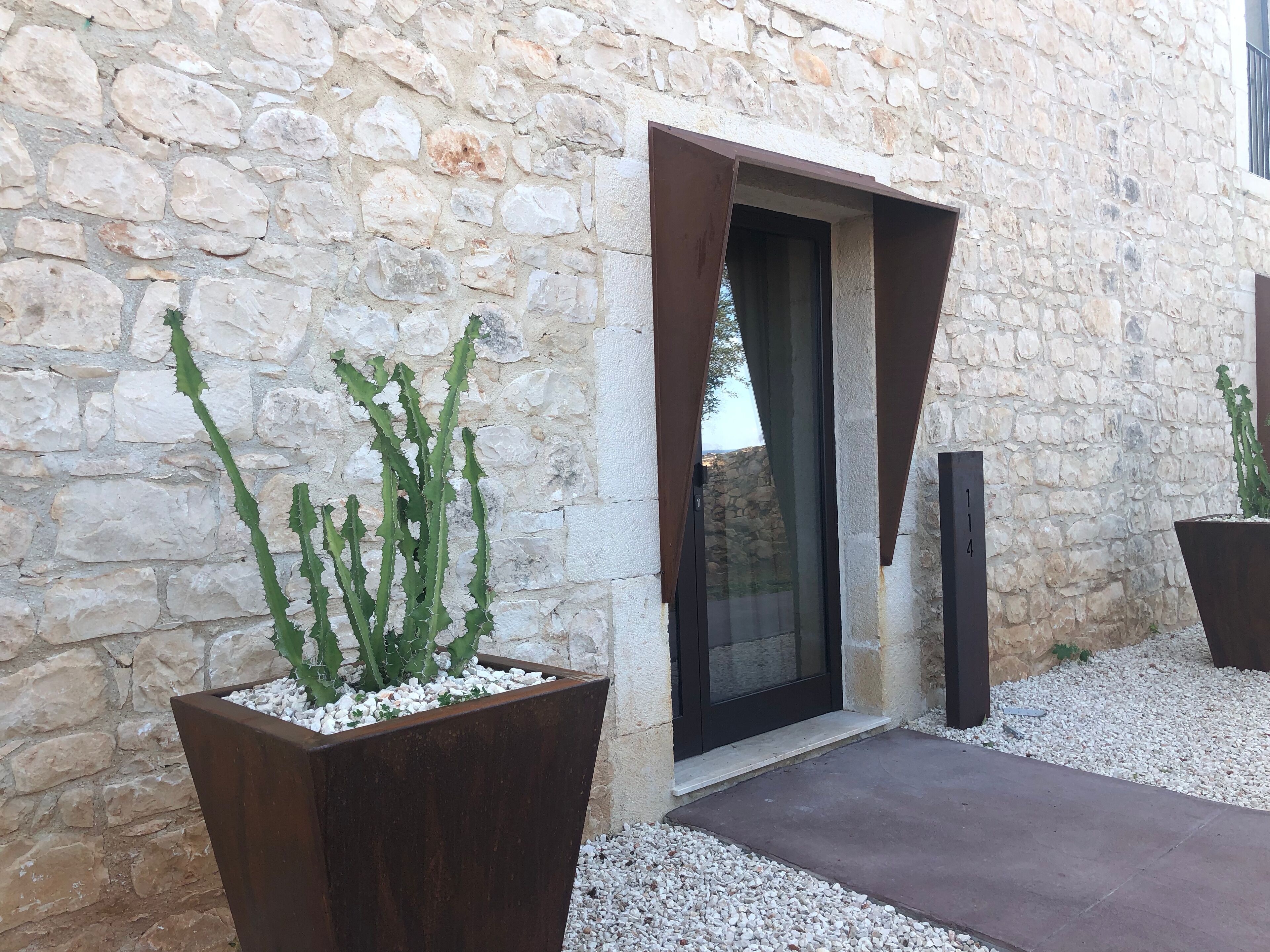 Un bâtiment en pierre doté d’une entrée moderne, de grands bacs à fleurs contenant des cactus et d’un chemin en béton.