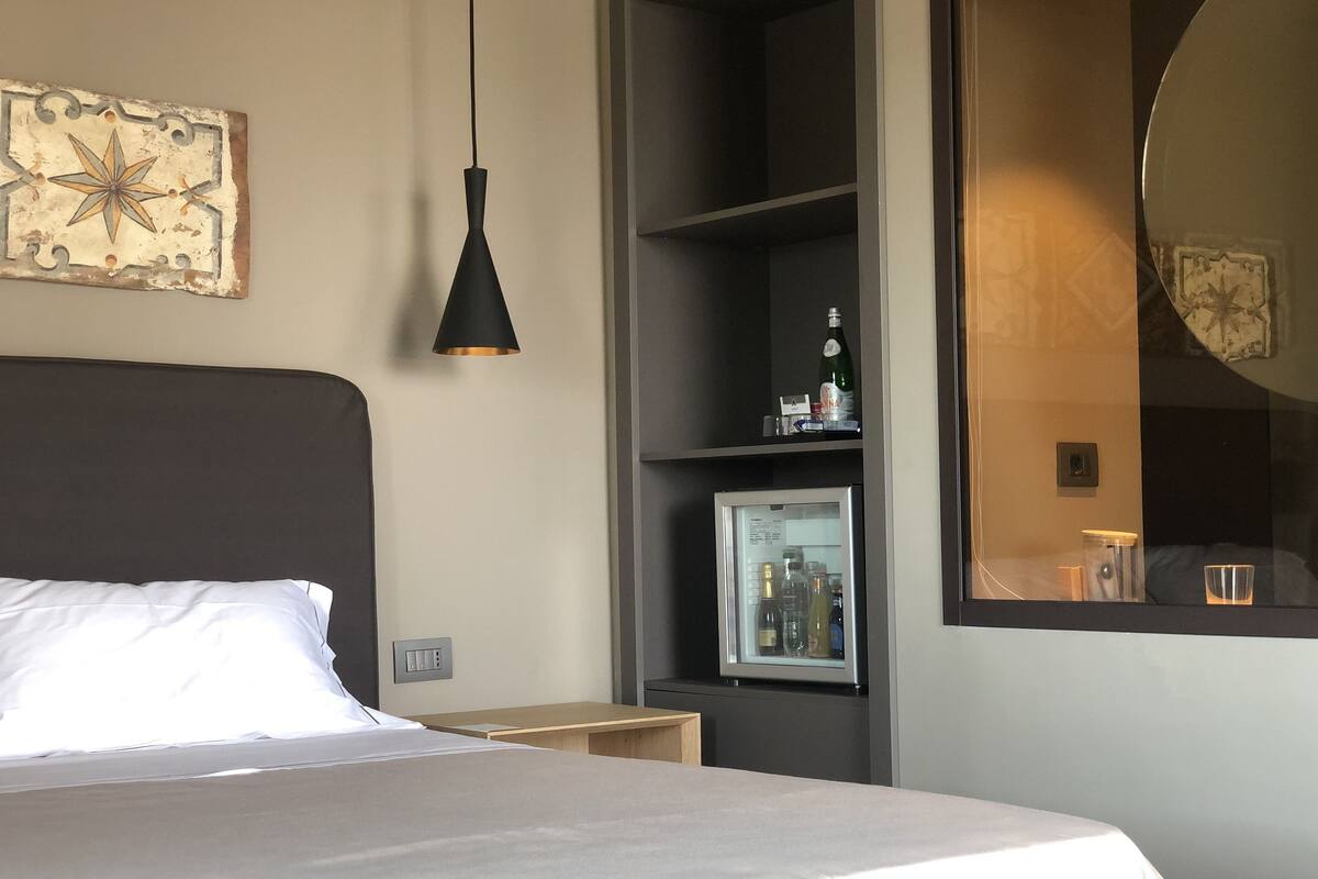 A hotel room with a bed, a black pendant light, a minibar, a mirror, and a framed picture on the wall.
