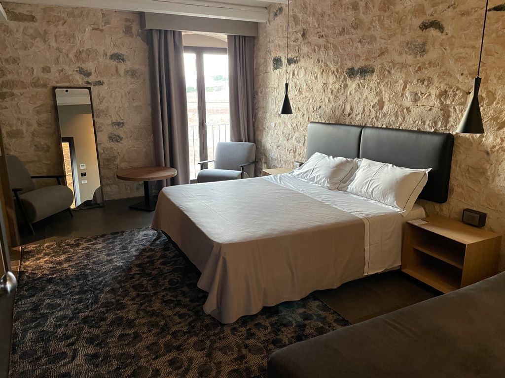 A hotel room with a large bed, a bedside table, a chair, a mirror, and a window with curtains.