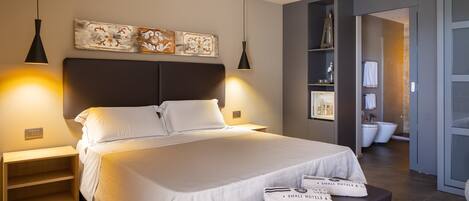 A modern hotel room with a large bed, bedside lamps, a nightstand, a small table with towels, and a bathroom visible through an open door.