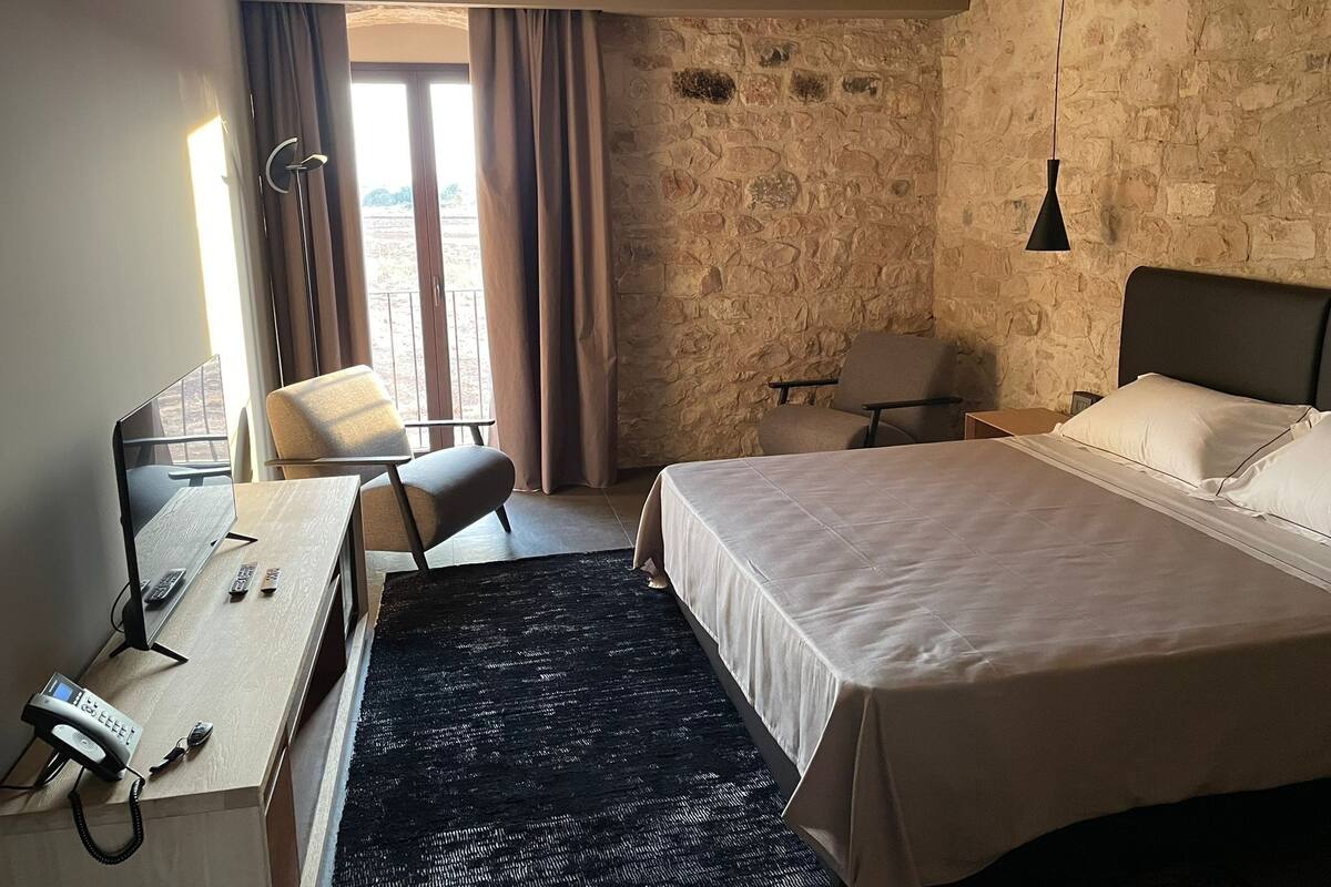 A hotel room with a bed, desk, chair, and a stone wall.