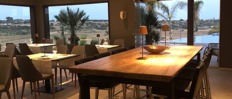 Un comedor moderno con una gran mesa de madera, varias sillas y dos lámparas de pie. Amplias ventanas ofrecen vistas al exterior.