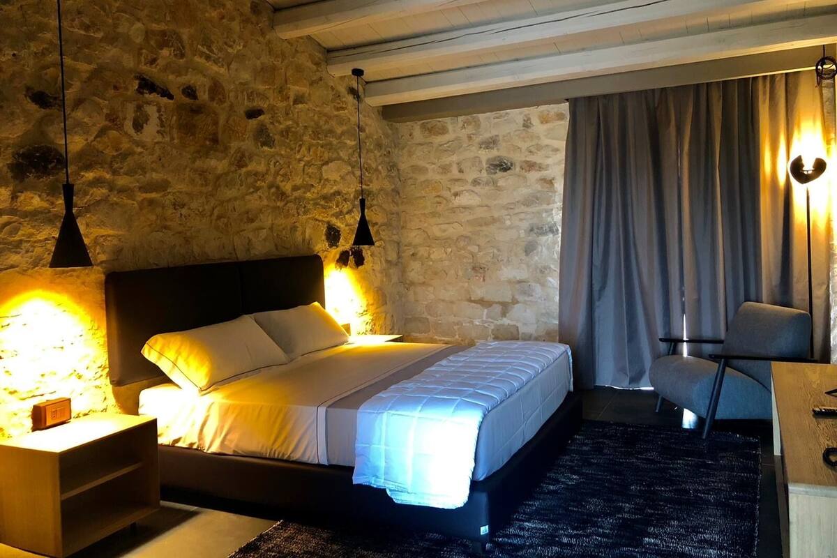 A bedroom with a stone wall, a bed with white bedding, a nightstand, a desk, and a chair.
