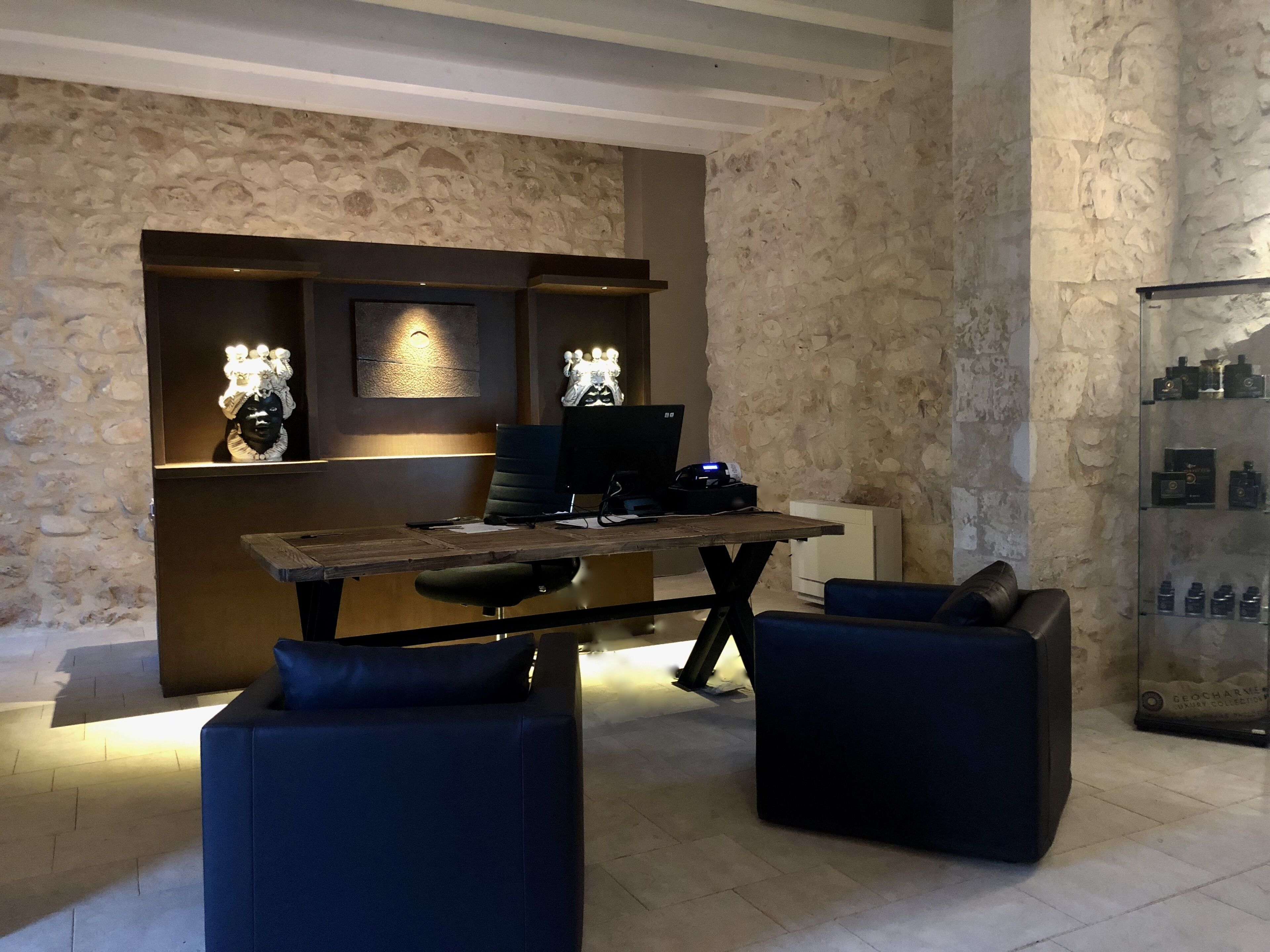 Un espacio de oficina moderno con una pared de piedra, un escritorio de madera, dos sillones azules y una estantería con objetos decorativos.