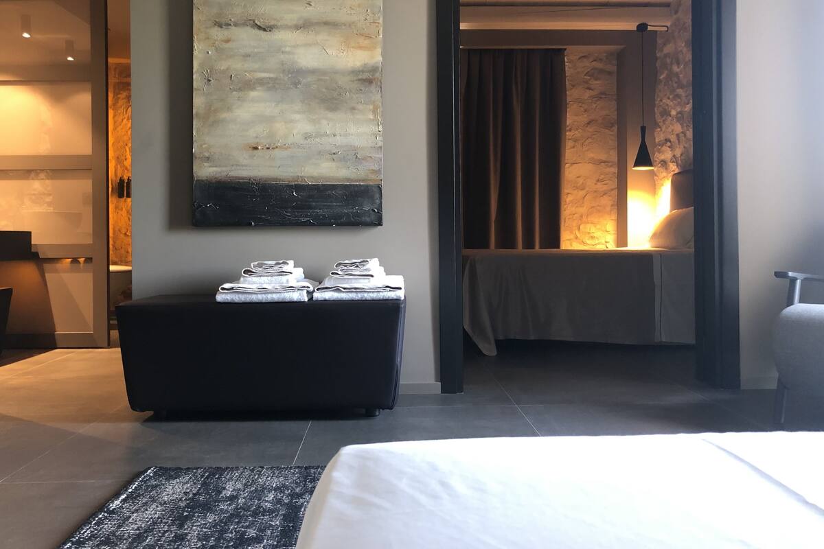 A modern hotel room with a large abstract painting, a neatly made bed, a black ottoman with towels, and a stone-textured wall.