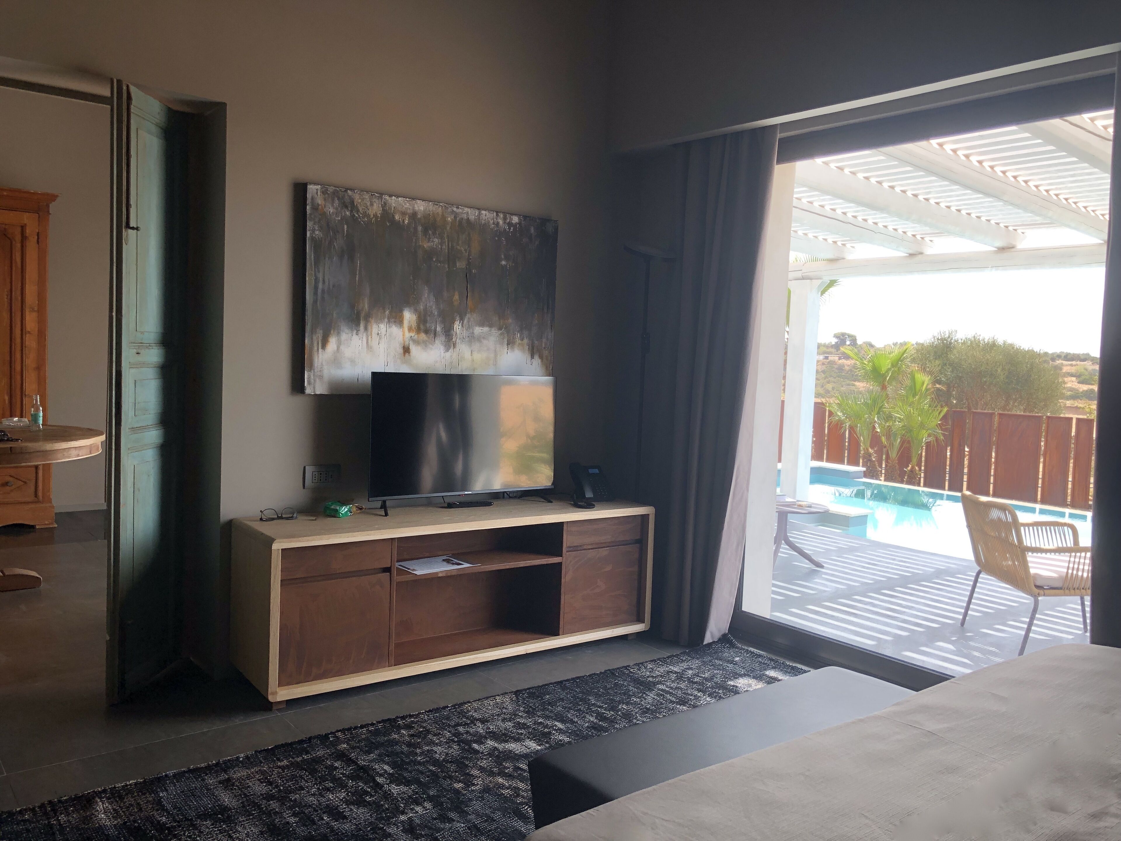 A modern hotel room with a large bed, a flat-screen TV on a wooden console, a painting on the wall, and a view of a swimming pool outside.