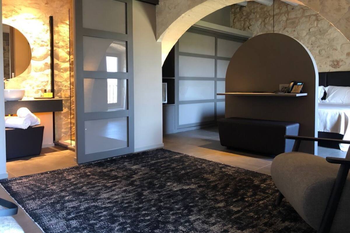 A modern hotel room with a stone wall, a round mirror, a black sofa, a small table, and a bed with white linens.