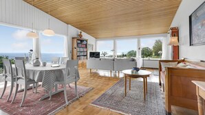 Een ruime woonkamer met een houten plafond, grote ramen met uitzicht op zee, een eethoek met een tafel en stoelen, en een gezellige zithoek met een bank en een klein tafeltje.