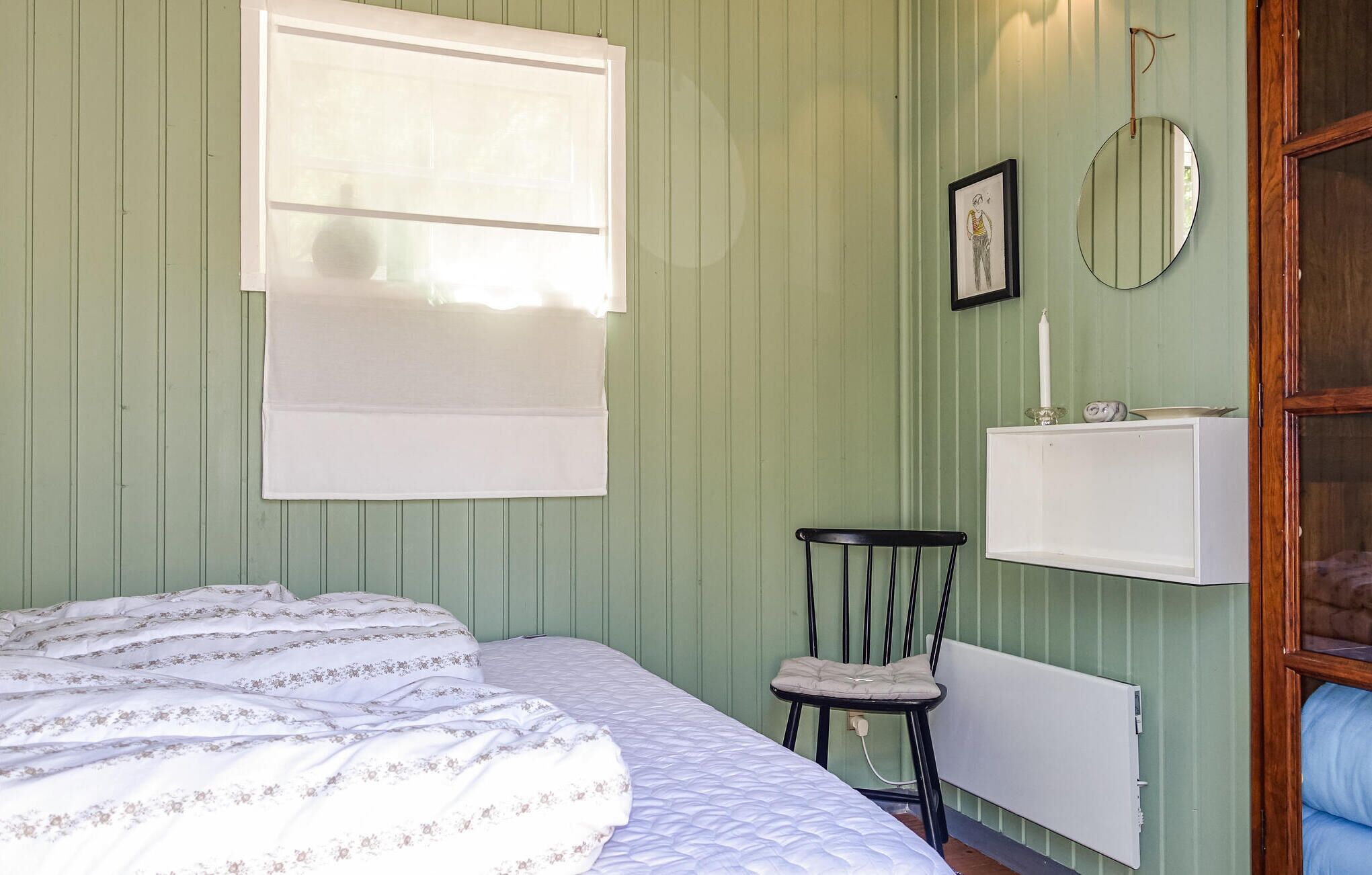 A room with a bed, a chair, a radiator, a mirror, and a shelf with a candle.