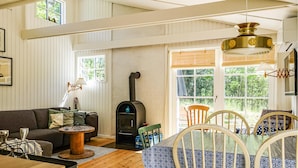 A cozy living room with a fireplace, wooden furniture, and large windows offering a view of greenery.
