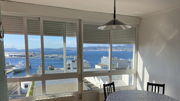 Une pièce dotée d’une grande fenêtre donnant sur une marina et un port, une table ronde recouverte d’une nappe à motifs floraux et un luminaire suspendu.