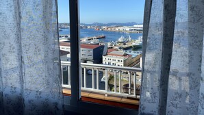 Une vue depuis une fenêtre ornée de rideaux à motifs floraux, donnant sur une marina avec des bateaux et des bâtiments.