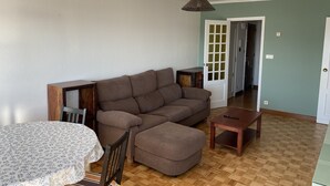 Un salon avec un canapé marron, une petite table en bois et une table à manger recouverte d’une nappe à motifs floraux.