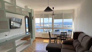 Un salon avec un canapé, une table et un téléviseur fixé au mur. De grandes fenêtres offrent une vue sur une marina.