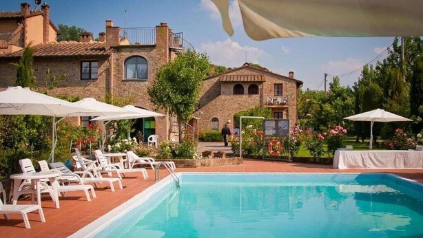 Zona de piscina con sillones de descanso y sombrillas, rodeada de edificios de piedra con balcones.