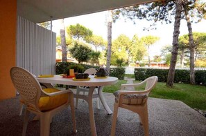 Een terras buiten met een ronde tafel en stoelen, een witte omheining en uitzicht op een tuin met bomen en een haag.