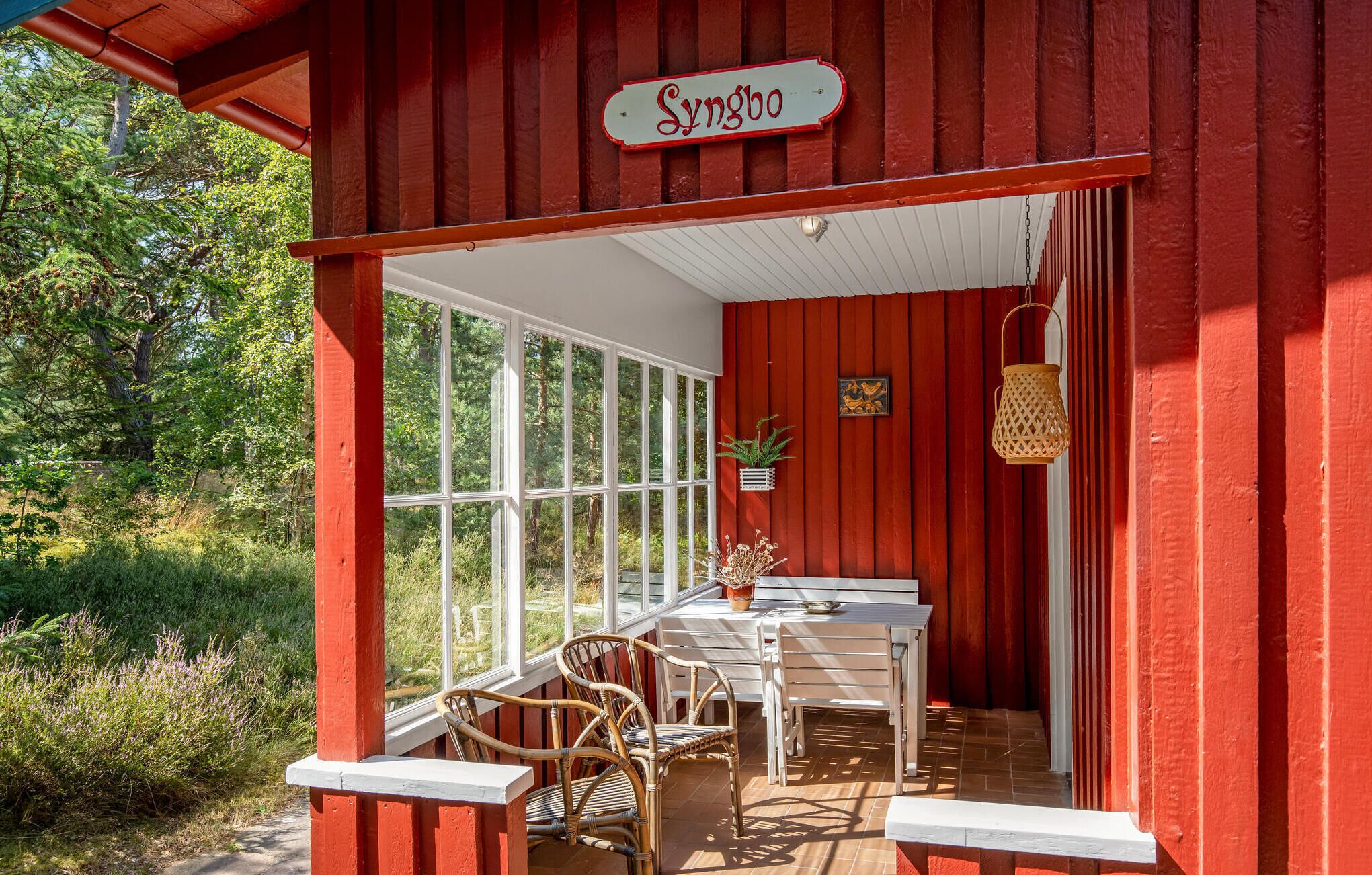 A red wooden building with a sign reading "Syngbo". Inside, there is a table and chairs set up near large windows, with a potted plant and a lantern on the wall.