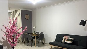 A modern living room with a black sofa, a dining table with chairs, a small potted plant, and a staircase in the background. - Spacious 3 bedroom townhouse with AC and lock up garage (Liverpool)