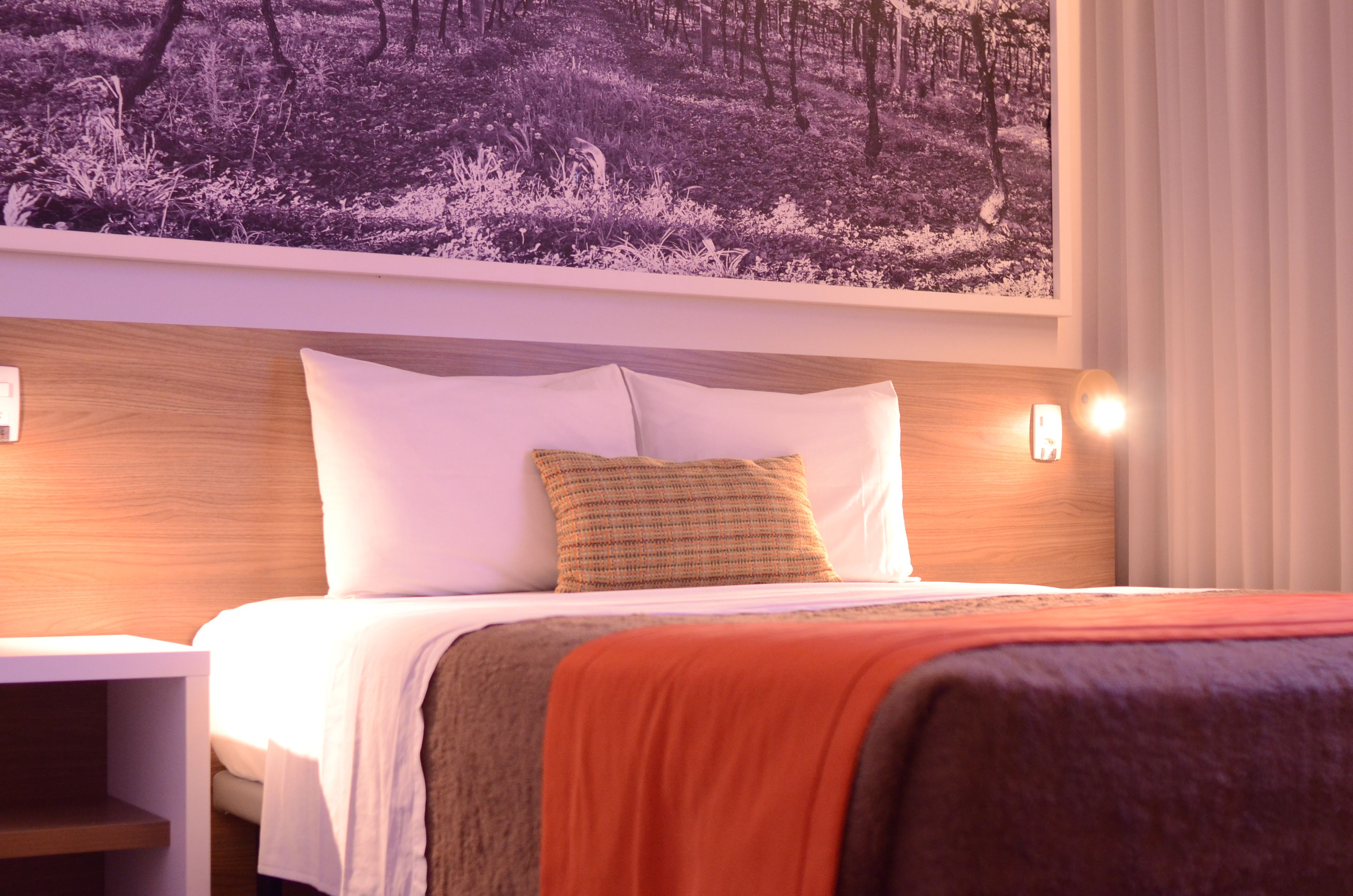 Une chambre d’hôtel avec un lit, une table de chevet, une lampe fixée au mur et un grand tableau encadré représentant une forêt.