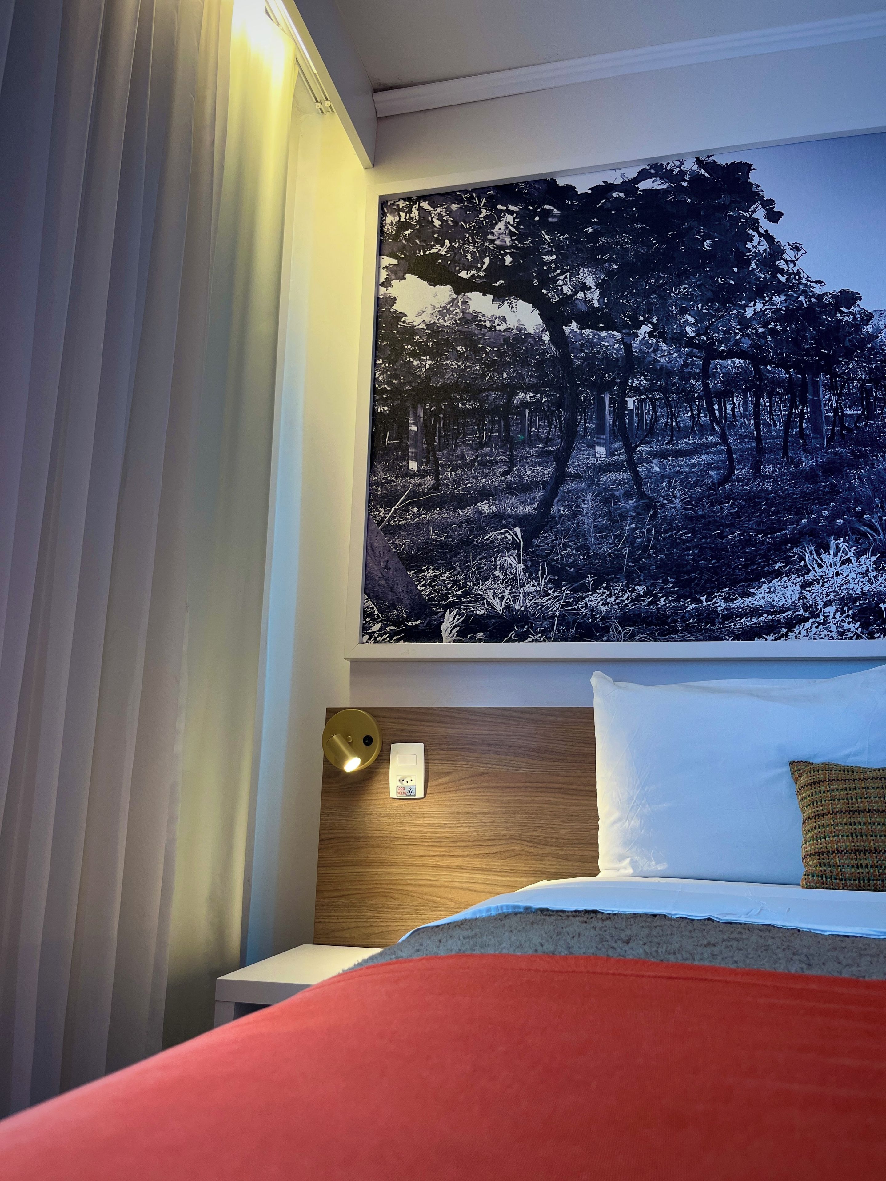 A hotel room with a bed, a nightstand, a wall-mounted lamp, and a large forest-themed artwork.