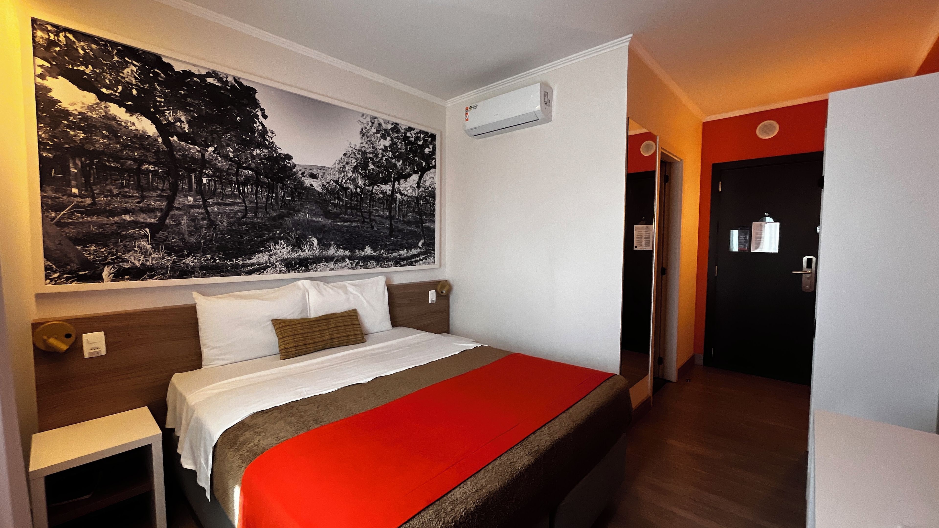 A hotel room with a bed, a large wall-mounted painting, an air conditioning unit, and a door leading to another room.