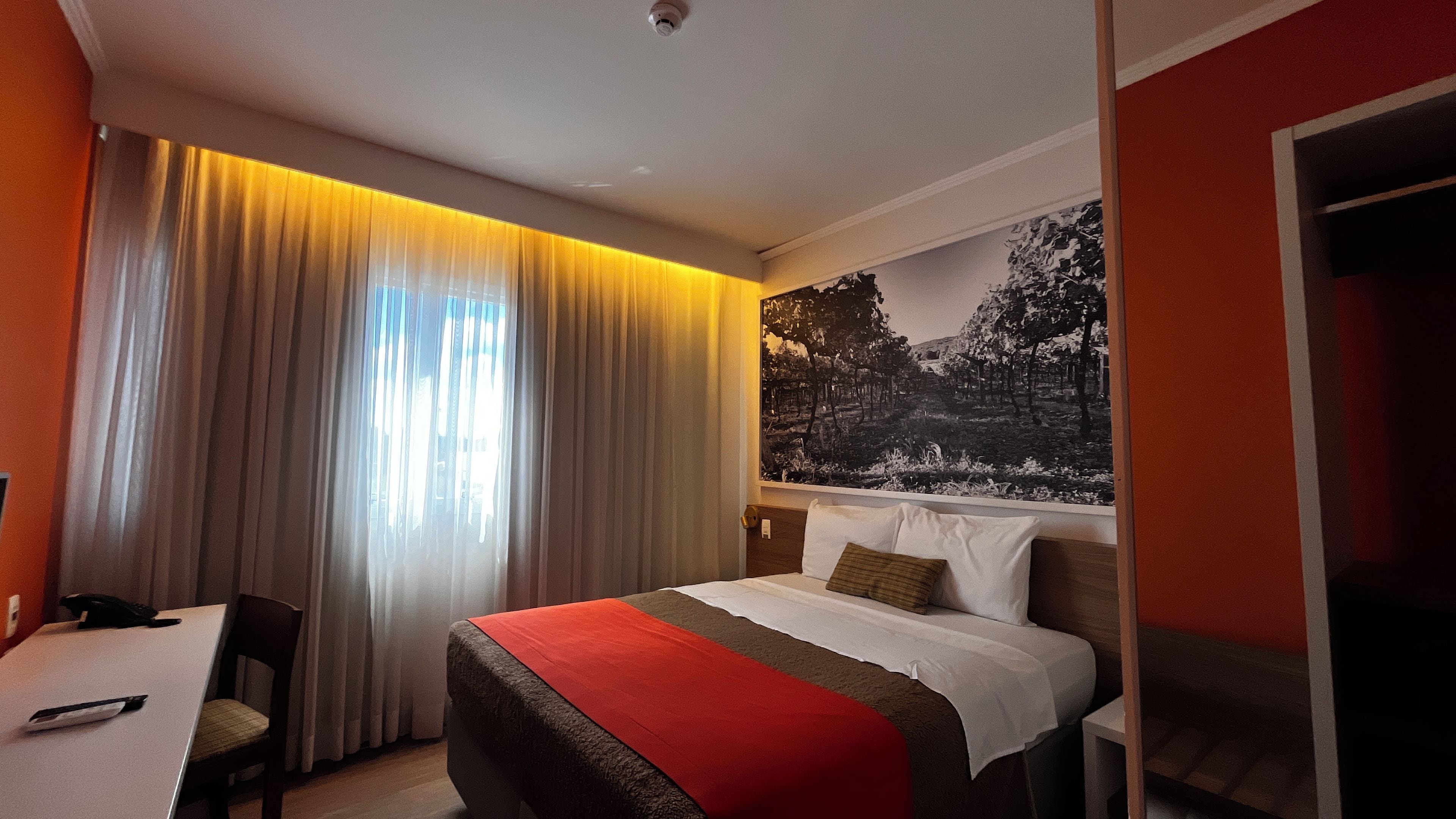 Una habitación de hotel con cama, escritorio, silla, un ventanal con cortinas y un cuadro colgado en la pared.