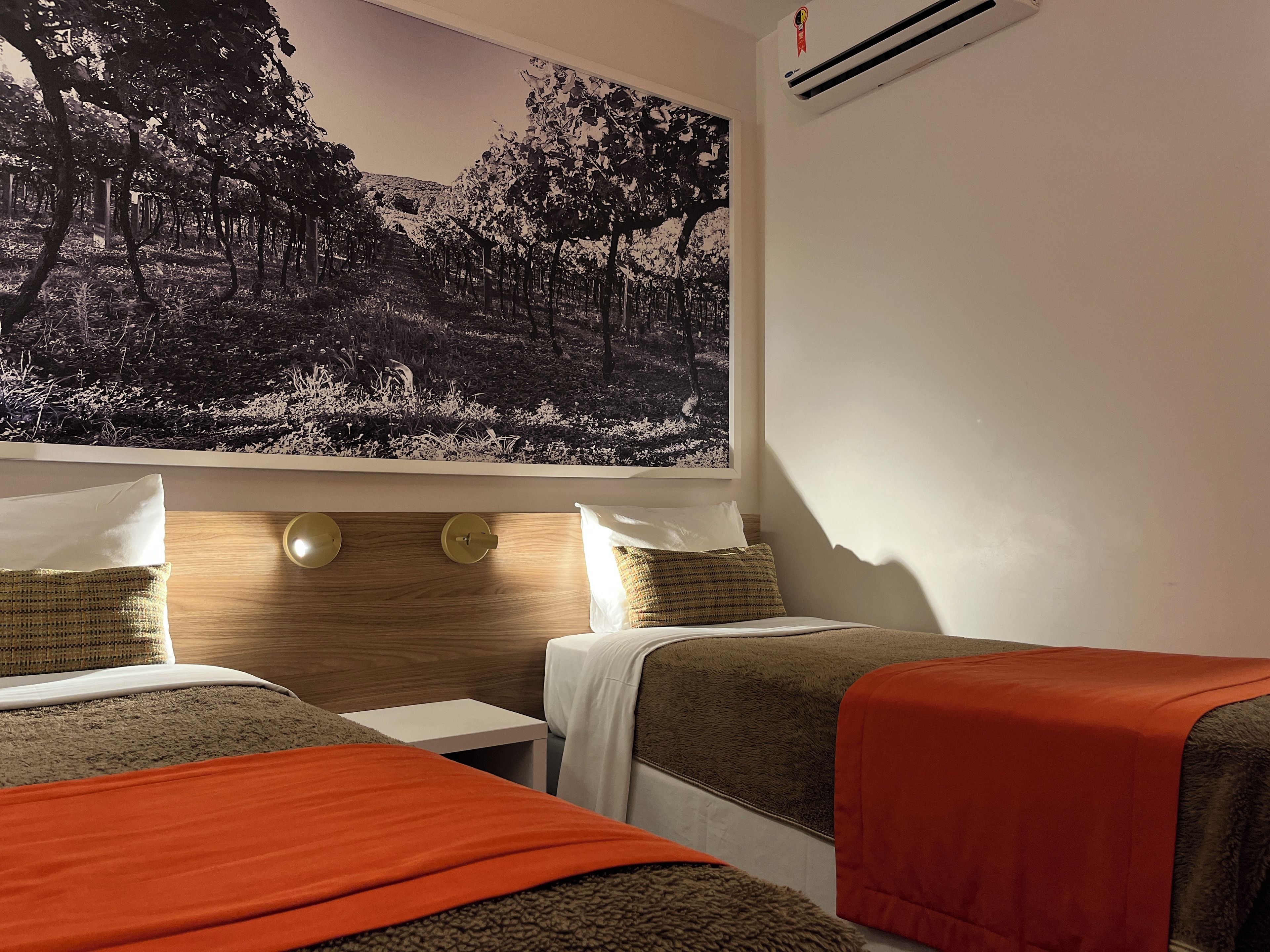 A hotel room with two beds, a nightstand, a wall-mounted air conditioner, and a large forest-themed painting.