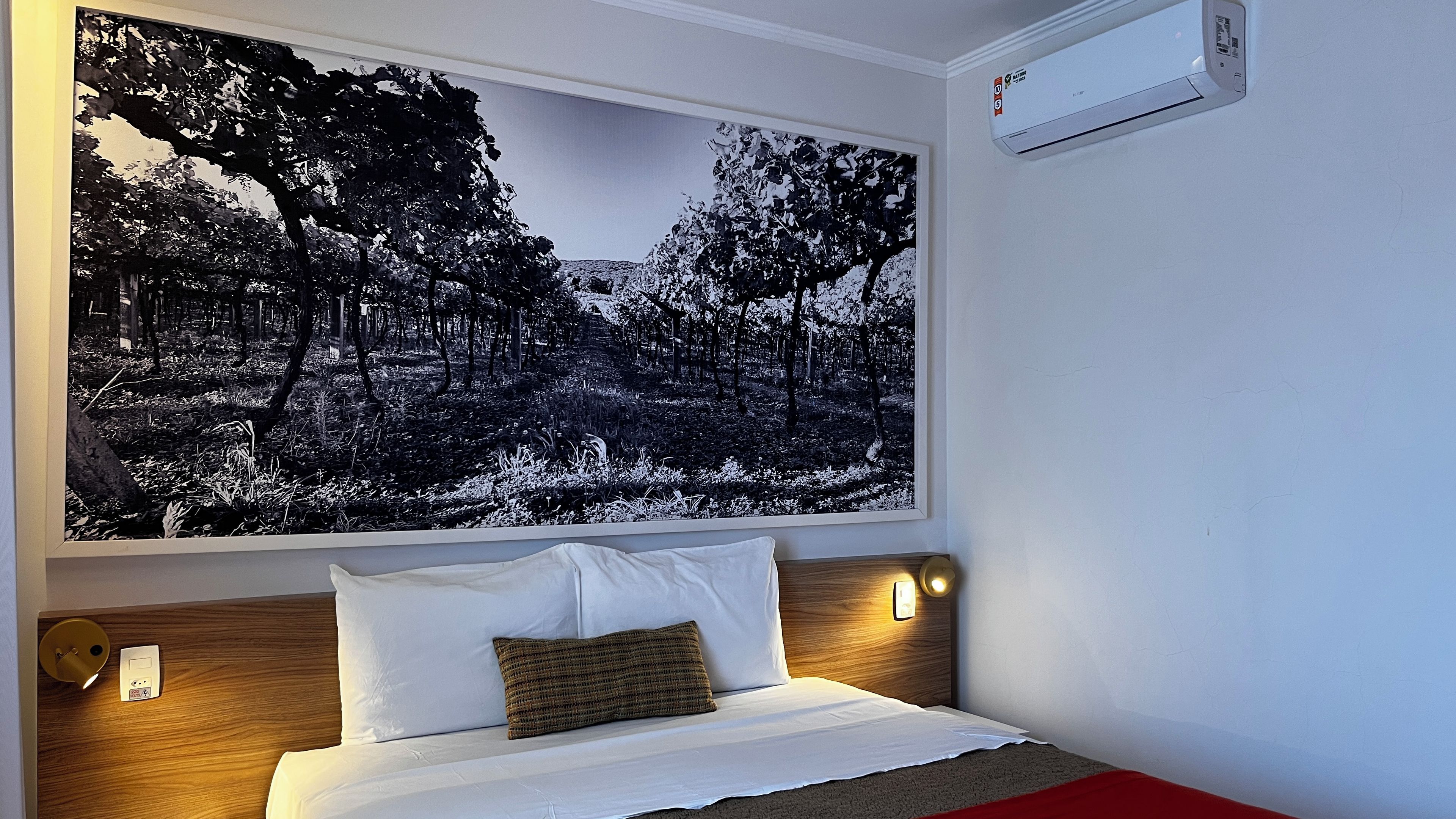 A hotel room with a bed, a wall-mounted air conditioner, and a large framed picture of a forest.