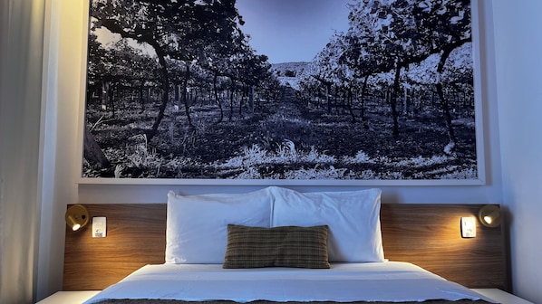 A hotel room with a large framed picture of a forest, a bed with white linens, a single brown pillow, and two wall-mounted light fixtures. - Partner Hotéis Caxias do Sul (Caxias do Sul)