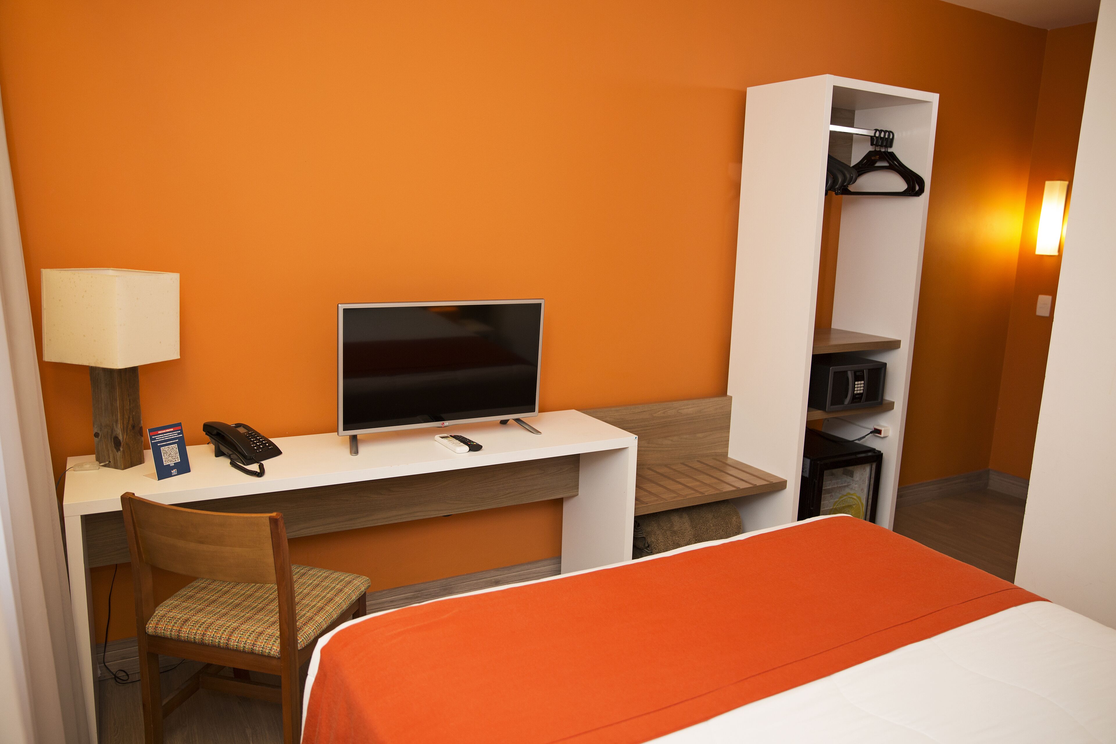 A hotel room with a bed, a desk with a TV, a chair, a lamp, and a wardrobe.