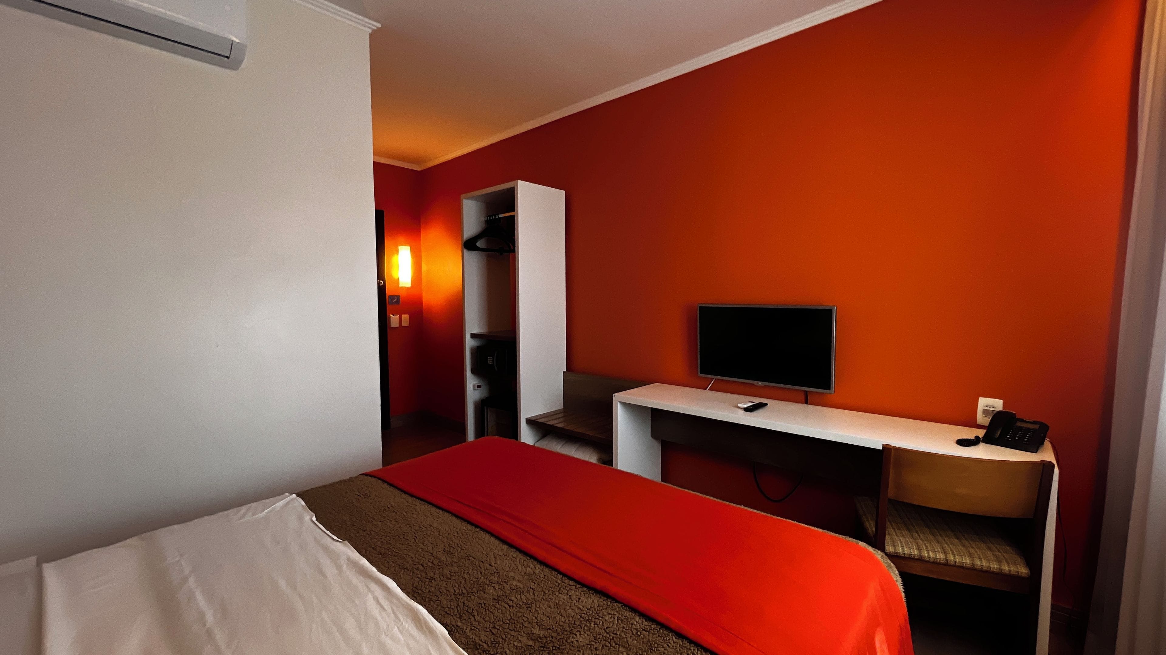 Quarto de hotel com cama, escrivaninha, televisão, cadeira e guarda-roupa.