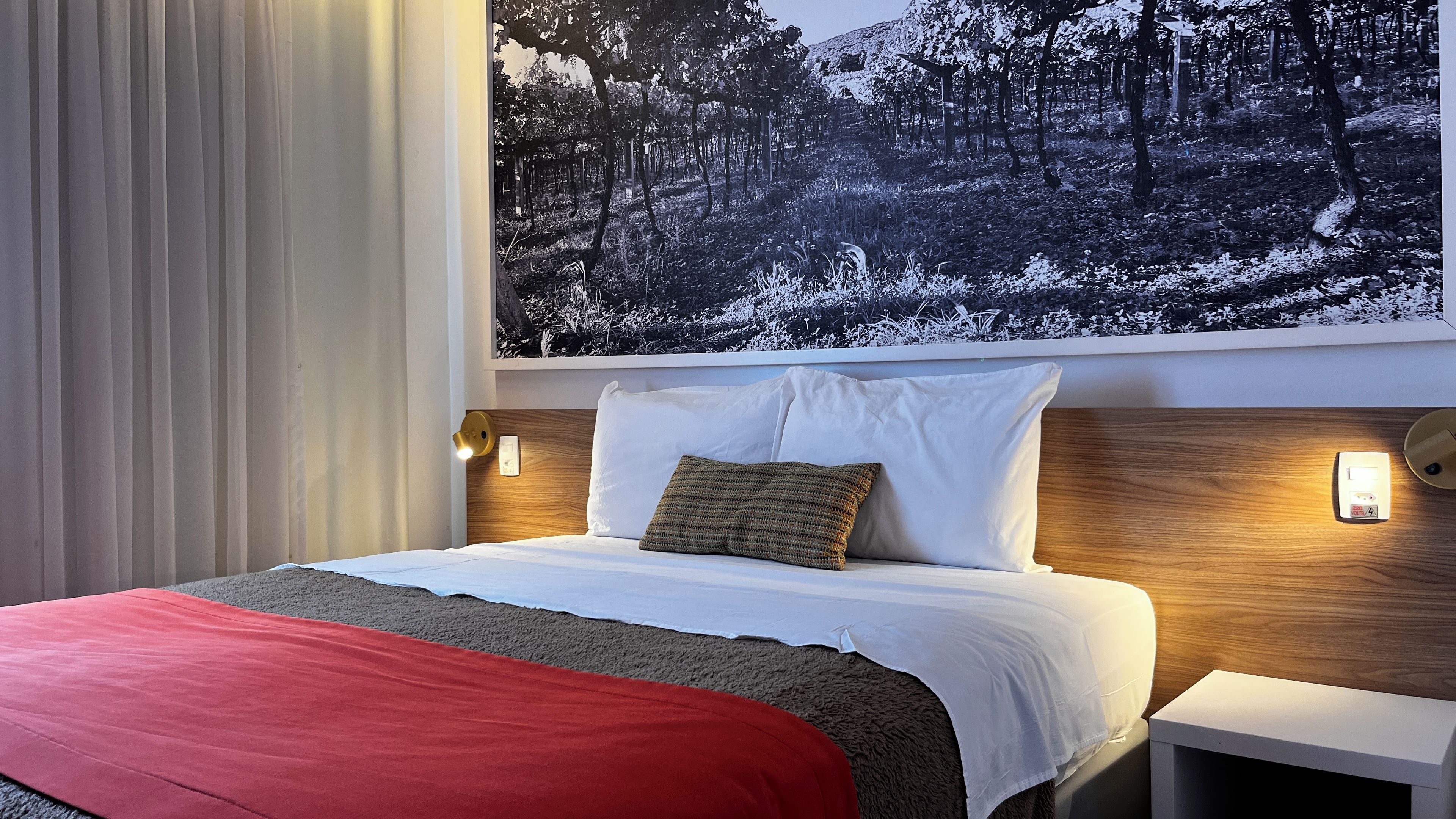 Una habitación de hotel con cama, mesita de noche, lámpara de pared y un cuadro grande con un paisaje de bosque.