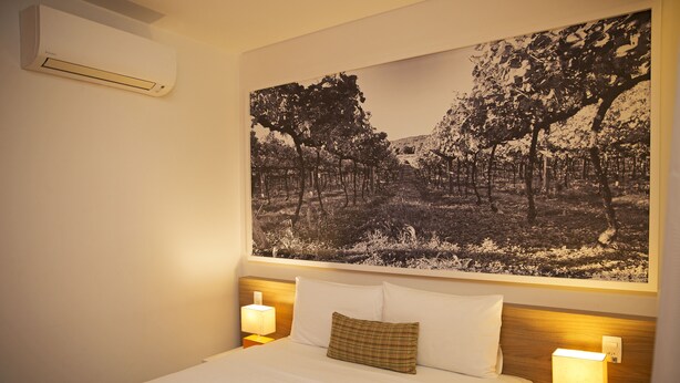 Een hotelkamer met een groot bed, twee bedlampen, een wandairconditioner en een groot zwart-wit foto van een bos boven het bed.