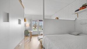 Et moderne soveværelse med en stor seng, et lille bord og en stol. Der er et vindue med gardiner, en hvid murstensvæg og et trægulv.