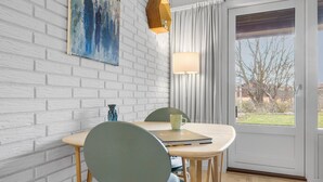 Et spiseområde med en hvid murstensvæg, et bord med stole, en lampe og udsigt gennem en glasdør.