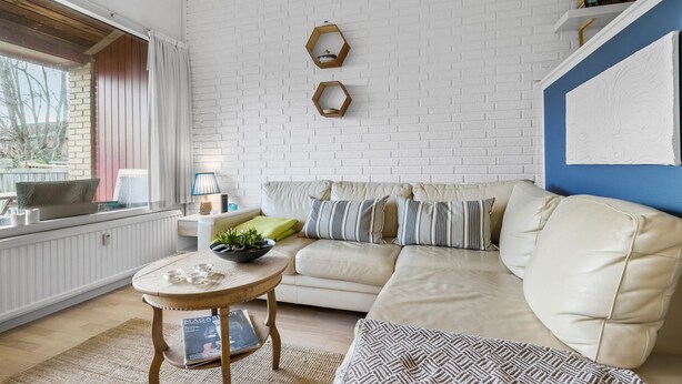 En stue med en hvid murstensvæg, en beige sofa med stribede puder, et rundt træbord med en skål planter og et vindue med gardiner.