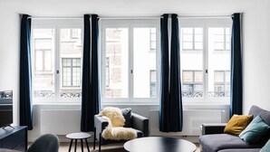 Een moderne woonkamer met een bank, fauteuil en salontafel, grote ramen met gordijnen en uitzicht op gebouwen.