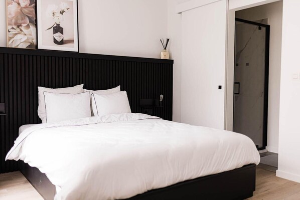 A modern bedroom with a black headboard, white bedding, and a door leading to a bathroom.