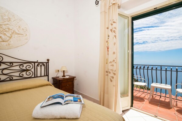 A bedroom with a bed, a nightstand, a lamp, and a view of a balcony with chairs. - Casa Lella (Furore)