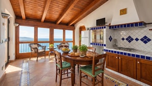 Una cocina con vigas de madera, una zona de comedor con sillas, una mesa con comida y vistas al mar.