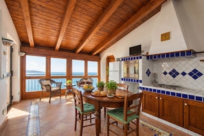 Una cocina con vigas de madera, una zona de comedor con sillas, una mesa con comida y vistas al mar.