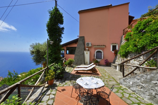 A coastal villa with a stone staircase, a lounge area with sun loungers, and a round table with chairs.